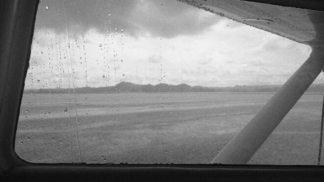 Rain-speckled aircraft window overlooking cloudy water and distant hills